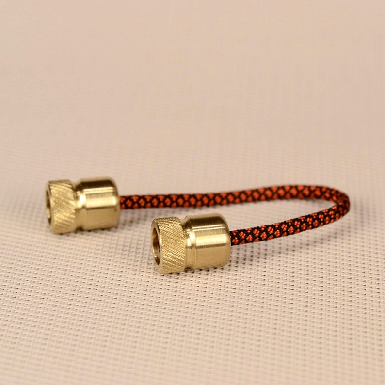 May include: A close-up of a fidget toy with two brass beads and a black and orange cord. The cord is woven with a diamond pattern.