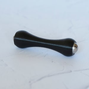 Op de afbeelding: Een zwart massagegereedschap in de vorm van een zandloper met een getextureerd oppervlak. Het heeft afgeronde uiteinden met zilveren metalen kogellagers. Ontworpen voor spierontspanning.