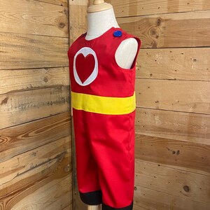 Peut inclure: Combinaison rouge avec un c&oelig;ur blanc et une ceinture jaune. La tenue sans manches a un bouton bleu sur l'&eacute;paule et une bordure noire aux chevilles. Le costume est pr&eacute;sent&eacute; sur un mannequin.