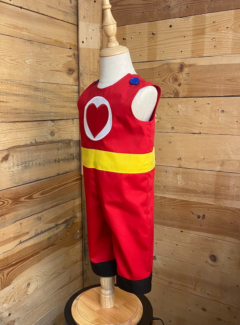 Peut inclure: Combinaison rouge avec un c&oelig;ur blanc et une ceinture jaune. La tenue sans manches a un bouton bleu sur l'&eacute;paule et une bordure noire aux chevilles. Le costume est pr&eacute;sent&eacute; sur un mannequin.