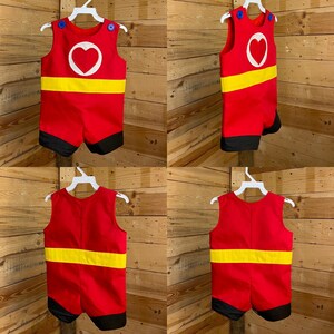 Puede incluir: Un pelele rojo con un dise&ntilde;o de coraz&oacute;n blanco, una franja amarilla y ribete negro. El atuendo sin mangas tiene detalles de botones azules y se muestra en una percha blanca. El pelele est&aacute; dise&ntilde;ado para ni&ntilde;os.