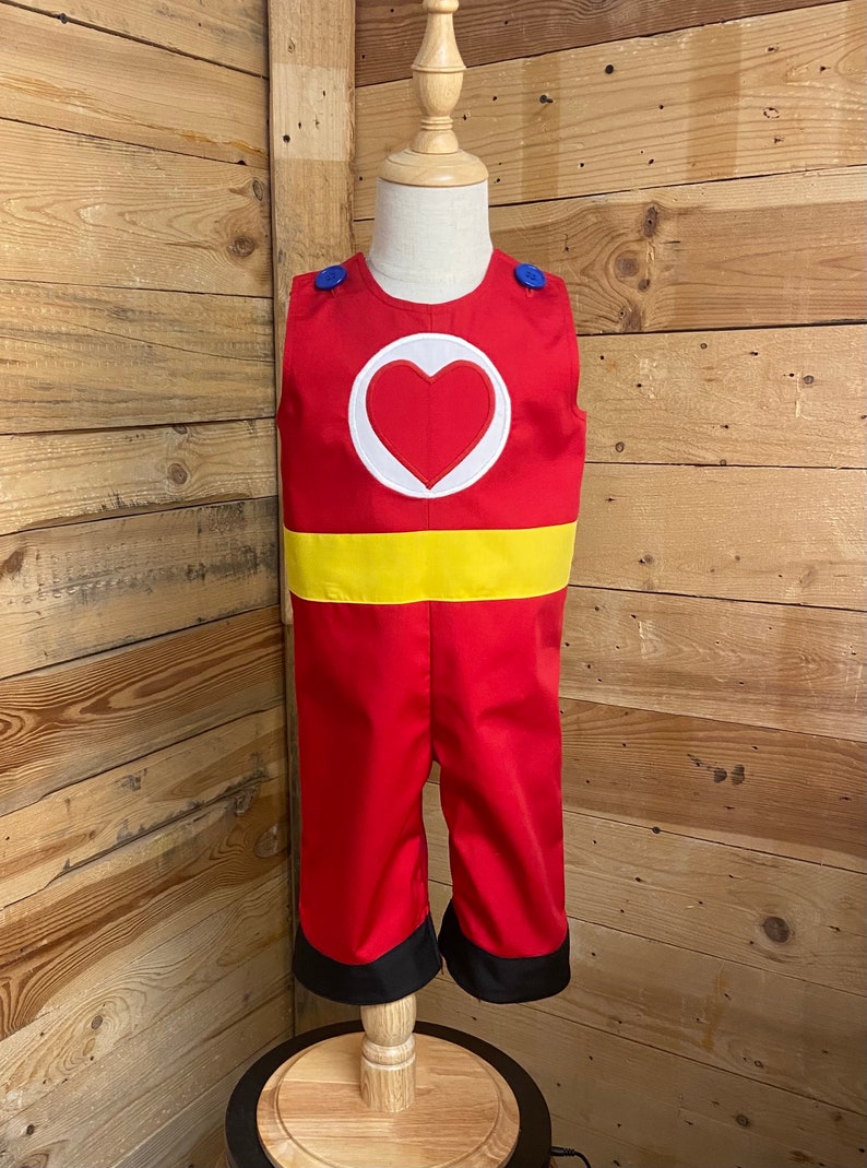 Peut inclure: Combinaison rouge pour enfant avec un c&oelig;ur blanc sur la poitrine, une ceinture jaune et des bordures noires aux jambes. Le costume a des boutons bleus sur les &eacute;paules. L'ensemble est pr&eacute;sent&eacute; sur un mannequin en bois.