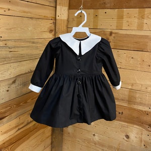 Pu&ograve; includere: Un abito nero con colletto bianco Peter Pan e maniche lunghe. L'abito ha una chiusura con bottoni sul davanti e una gonna a pieghe.