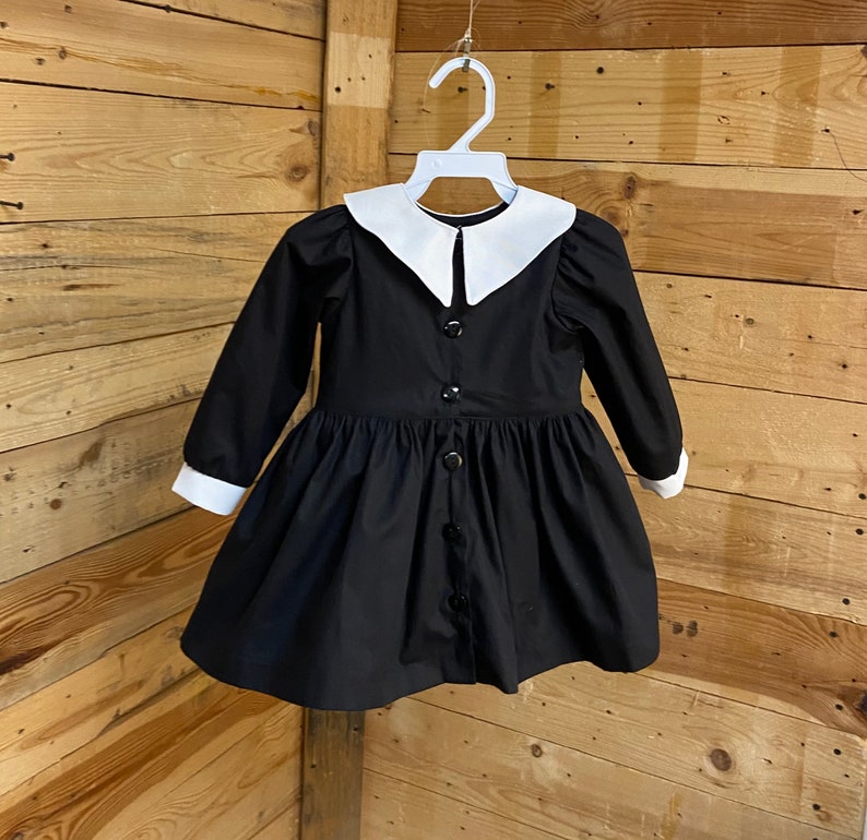 Pu&ograve; includere: Un abito nero con colletto bianco Peter Pan e maniche lunghe. L'abito ha una chiusura con bottoni sul davanti e una gonna a pieghe.