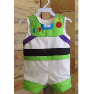 Puede incluir: Un pelele con el tema de Buzz Lightyear para beb&eacute; o ni&ntilde;o peque&ntilde;o, con un cuerpo blanco con detalles verdes y negros. La parte superior tiene un cuello y detalles verdes, incluyendo un c&iacute;rculo rojo y un rect&aacute;ngulo azul. El conjunto es sin mangas y tiene pantalones cortos verdes.