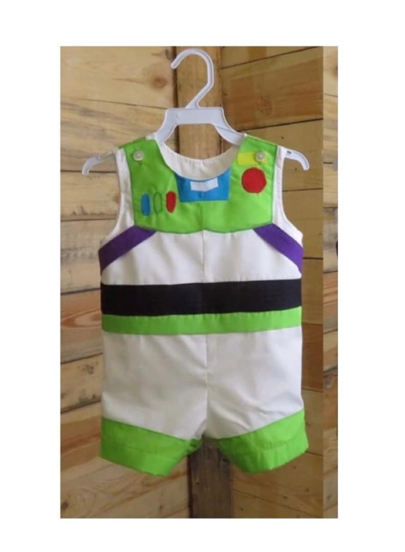 Puede incluir: Un pelele con el tema de Buzz Lightyear para beb&eacute; o ni&ntilde;o peque&ntilde;o, con un cuerpo blanco con detalles verdes y negros. La parte superior tiene un cuello y detalles verdes, incluyendo un c&iacute;rculo rojo y un rect&aacute;ngulo azul. El conjunto es sin mangas y tiene pantalones cortos verdes.