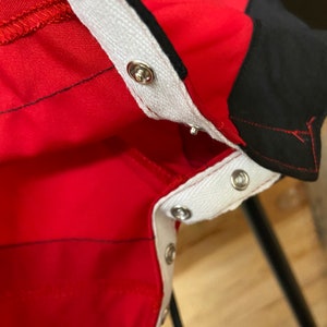 Puede incluir: Primer plano de una prenda roja con ribete blanco y cierres de presi&oacute;n plateados. La prenda tiene detalles en negro y costuras negras. El ribete blanco est&aacute; cosido a la tela roja. La imagen es un primer plano de los detalles de la prenda.