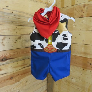 May include: A cowboy-themed costume set for a child, featuring a blue romper, a vest with a cow-print pattern, a red neckerchief, and a gold star badge. The outfit is designed to resemble a classic cowboy character.
