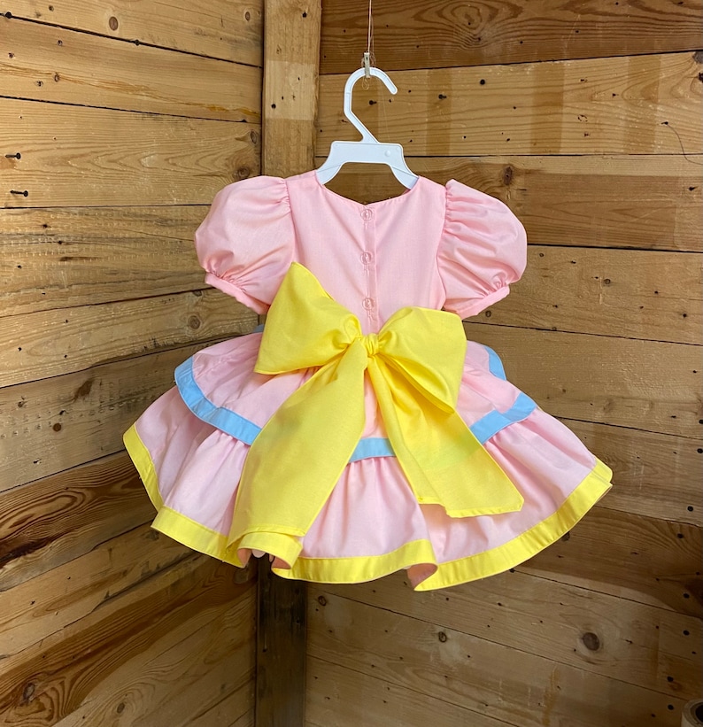 May include: A pink baby dress with a large yellow bow, short puffed sleeves, and a tiered skirt with blue and yellow accents. The dress is hanging on a white hanger against a wooden background.