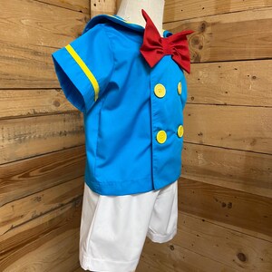 May include: A blue and white sailor costume for a child. The costume includes a short-sleeved shirt with yellow trim, a red bow tie, and white shorts.