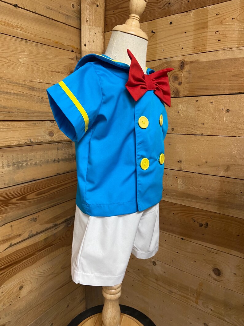 May include: A blue and white sailor costume for a child. The costume includes a short-sleeved shirt with yellow trim, a red bow tie, and white shorts.