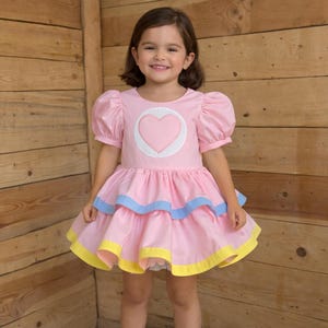 May include: Pink dress with a heart design. The dress features short, puffy sleeves, a layered skirt with blue and yellow trim, and a heart applique on the bodice. The dress is designed for a child.