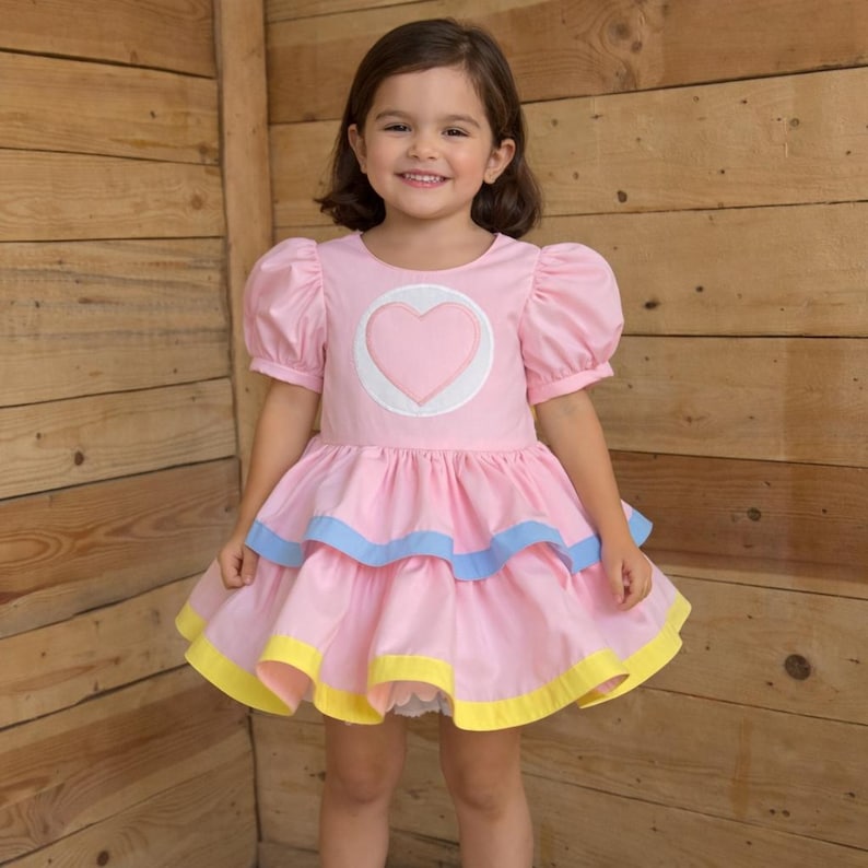 May include: Pink dress with a heart design. The dress features short, puffy sleeves, a layered skirt with blue and yellow trim, and a heart applique on the bodice. The dress is designed for a child.