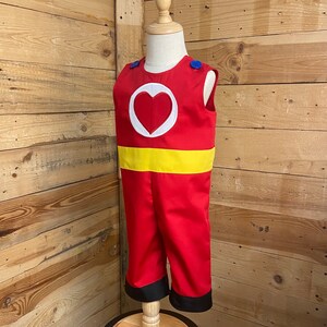 Peut inclure: Un costume de combinaison rouge avec un embl&egrave;me de c&oelig;ur blanc et une ceinture jaune. La tenue sans manches a des boutons bleus sur les &eacute;paules et une bordure noire aux chevilles. Le costume est pr&eacute;sent&eacute; sur un mannequin.