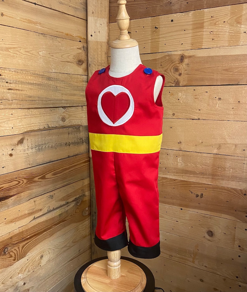 Peut inclure: Un costume de combinaison rouge avec un embl&egrave;me de c&oelig;ur blanc et une ceinture jaune. La tenue sans manches a des boutons bleus sur les &eacute;paules et une bordure noire aux chevilles. Le costume est pr&eacute;sent&eacute; sur un mannequin.