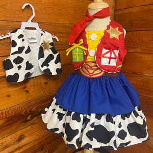 May include: A child's costume featuring a yellow shirt with white buttons, a red vest with a gold star, and a blue and cow-print skirt. A matching cow-print vest hangs on a hanger. The costume is displayed on a wooden mannequin.