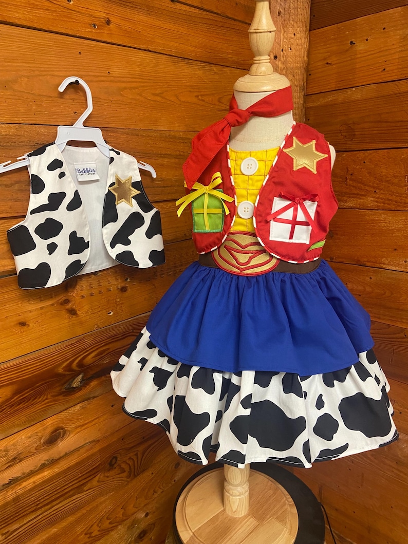 May include: A child's costume featuring a yellow shirt with white buttons, a red vest with a gold star, and a blue and cow-print skirt. A matching cow-print vest hangs on a hanger. The costume is displayed on a wooden mannequin.