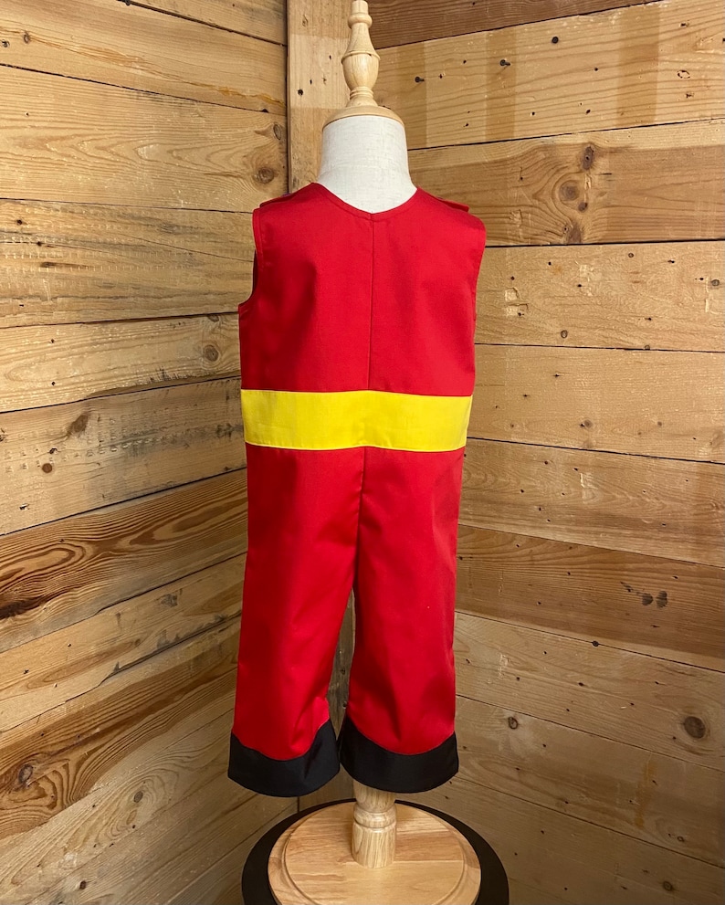 Peut inclure: Combinaison rouge avec une ceinture jaune et des poignets noirs, pr&eacute;sent&eacute;e sur un mannequin en bois. La tenue sans manches a un col en V. L'arri&egrave;re-plan est un mur en bois.