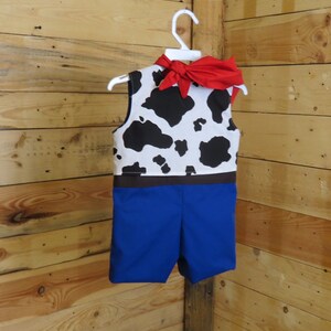 May include: A child's costume resembling a cowboy outfit, featuring a vest with a black and white cow pattern, blue shorts, and a red neckerchief tied in a bow. The outfit is hanging on a white hanger against a wooden background.