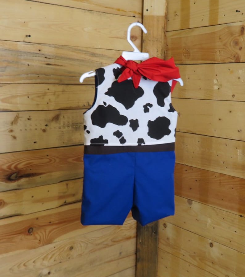 May include: A child's costume resembling a cowboy outfit, featuring a vest with a black and white cow pattern, blue shorts, and a red neckerchief tied in a bow. The outfit is hanging on a white hanger against a wooden background.