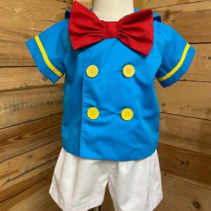 May include: A blue and white sailor costume for a child. The costume includes a short-sleeved shirt with yellow trim, a red bow tie, and white shorts.