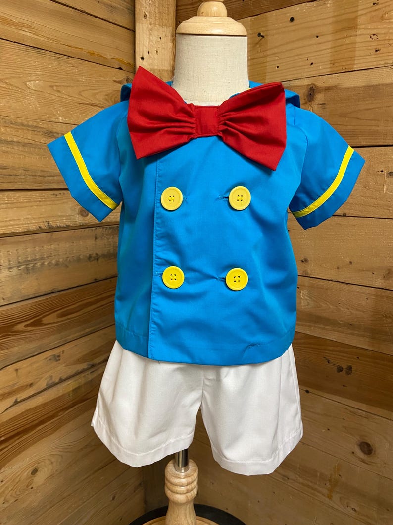 May include: A blue and white sailor costume for a child. The costume includes a short-sleeved shirt with yellow trim, a red bow tie, and white shorts.