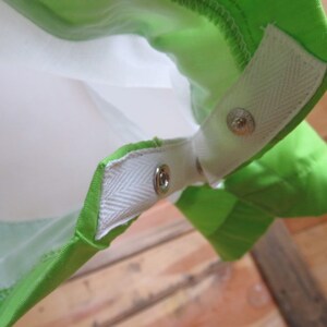 May include: Close-up of a bright green fabric with white accents and metal snap closures. The fabric appears to be part of a larger item, possibly a bag or garment, with a white interior visible.