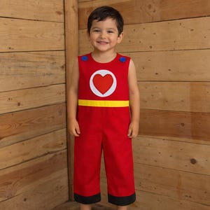 Peut inclure: Combinaison rouge pour enfant avec un c&oelig;ur rouge dans un cercle blanc sur la poitrine. Une bande jaune orne la taille et des bordures noires les bas de jambes. Des boutons bleus sont sur les &eacute;paules.