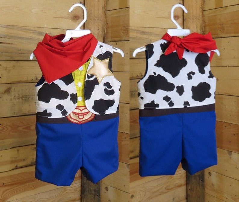 May include: A two-piece costume resembling a cowboy outfit, featuring a vest with a cow-print design, a yellow shirt, and blue shorts. The vest has a sheriff's star and a red bandana. The costume is hanging on a white hanger.