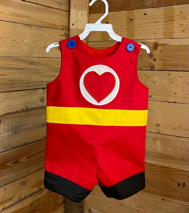 Puede incluir: Romper rojo con cinturilla amarilla y una aplicaci&oacute;n de coraz&oacute;n con borde blanco. El romper tiene botones azules en los hombros y ribete negro en los pantalones cortos. La prenda est&aacute; colgada de una percha blanca sobre un fondo de madera.