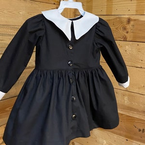 Pu&ograve; includere: Un abito nero con colletto bianco Peter Pan e maniche lunghe. L'abito ha una chiusura con bottoni sul davanti e una gonna a pieghe.