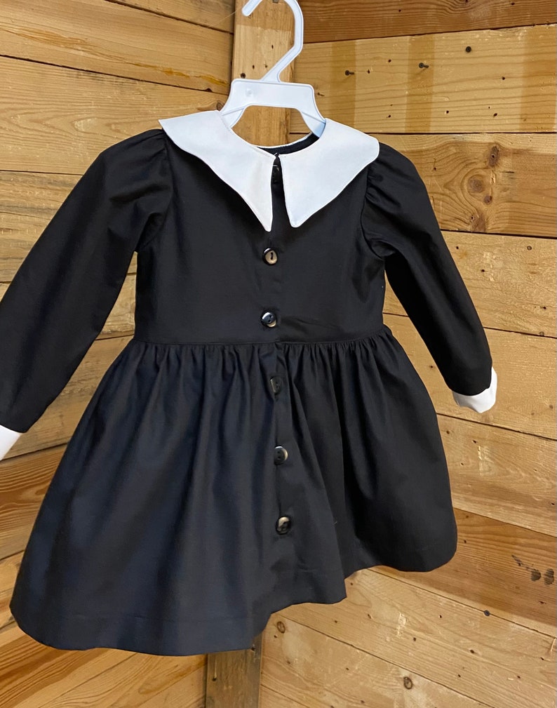 Pu&ograve; includere: Un abito nero con colletto bianco Peter Pan e maniche lunghe. L'abito ha una chiusura con bottoni sul davanti e una gonna a pieghe.