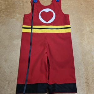Peut inclure: Combinaison rouge avec un c&oelig;ur blanc dans un cercle sur la poitrine. La combinaison a une bande jaune au milieu et une bordure noire en bas. Des rubans noirs ornent le devant et des boutons bleus fixent les bretelles.