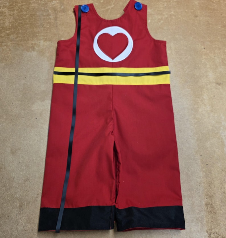 Peut inclure: Combinaison rouge avec un c&oelig;ur blanc dans un cercle sur la poitrine. La combinaison a une bande jaune au milieu et une bordure noire en bas. Des rubans noirs ornent le devant et des boutons bleus fixent les bretelles.