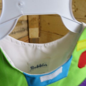 Puede incluir: Un vestido colorido con cuello blanco y una etiqueta que dice "Bubbles". El vestido tiene un dise&ntilde;o verde, azul, rojo y amarillo. El vestido est&aacute; colgado en una percha blanca.