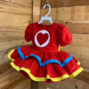 Pode incluir: Um vestido vermelho com um cora&ccedil;&atilde;o branco na frente e acabamento amarelo e azul ao redor da bainha. O vestido tem uma manga curta bufante.