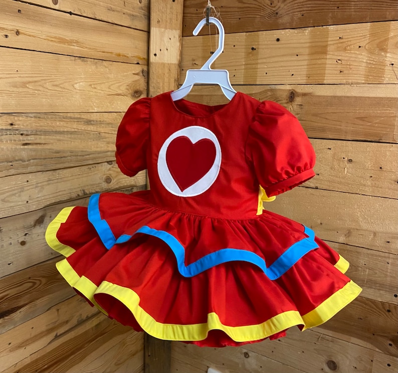 Pode incluir: Um vestido vermelho com um cora&ccedil;&atilde;o branco na frente e acabamento amarelo e azul ao redor da bainha. O vestido tem uma manga curta bufante.