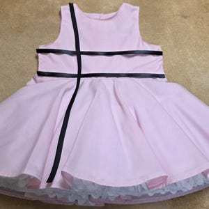 May include: A light pink dress with a full skirt and a black ribbon detail. The dress has a sleeveless bodice and a white lace trim at the hem. The dress is designed for a child.