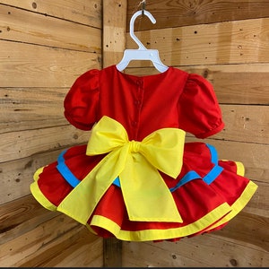 Pode incluir: Vestido vermelho com um la&ccedil;o amarelo e acabamento azul. O vestido tem uma saia com babados e mangas curtas.