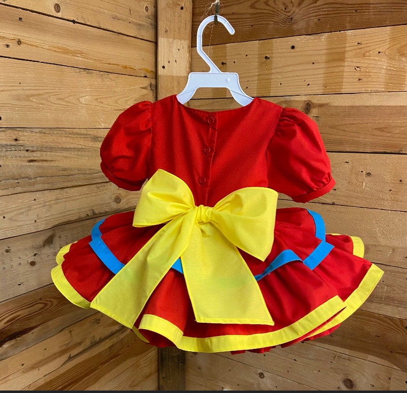 Pode incluir: Vestido vermelho com um la&ccedil;o amarelo e acabamento azul. O vestido tem uma saia com babados e mangas curtas.