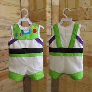 Puede incluir: Un disfraz de Buzz Lightyear blanco y verde para beb&eacute;, con cintur&oacute;n negro, detalles morados y cuello verde. El disfraz se muestra en una percha blanca sobre un fondo de madera. Se ven la parte delantera y trasera del disfraz.