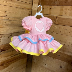 May include: A pink dress with a heart-shaped design on the bodice and layered skirt. The dress features short, puffed sleeves and is trimmed with blue and yellow accents. It hangs on a white hanger.