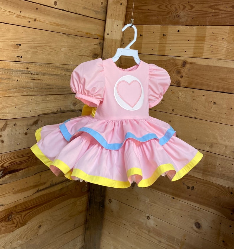 May include: A pink dress with a heart-shaped design on the bodice and layered skirt. The dress features short, puffed sleeves and is trimmed with blue and yellow accents. It hangs on a white hanger.