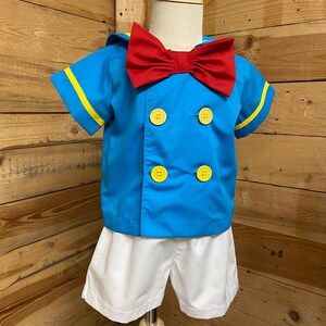 May include: A blue and white sailor costume for a child. The shirt has yellow trim and red bow tie. The shorts are white.