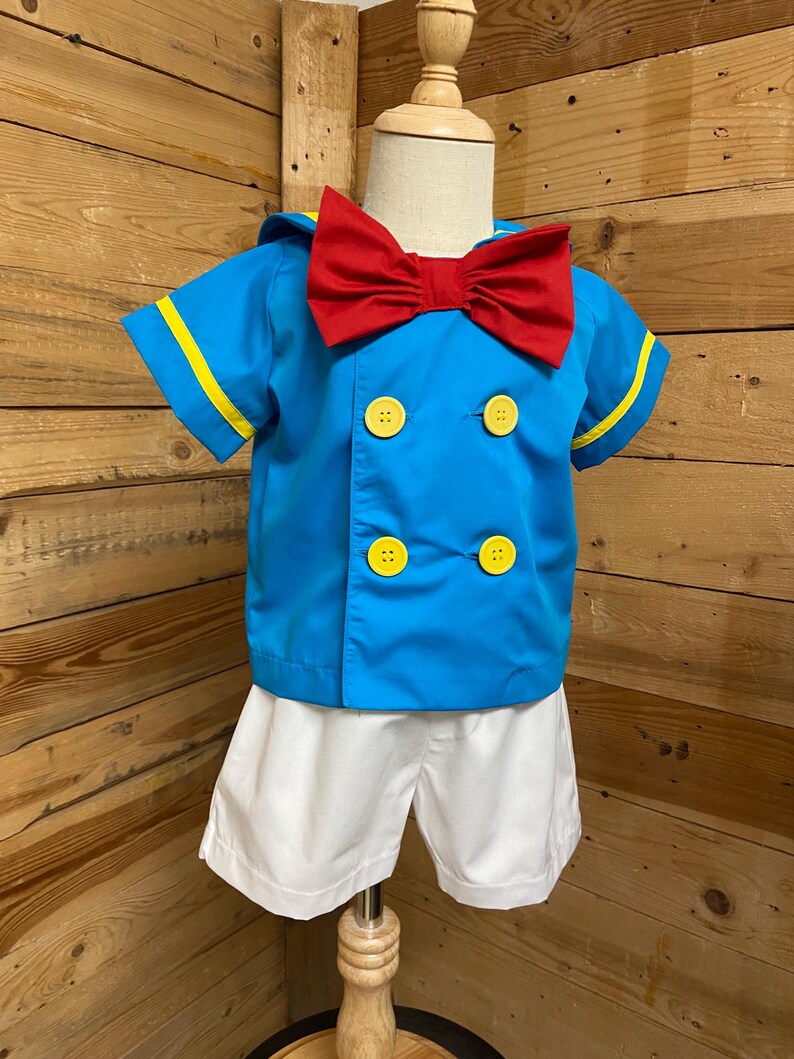 May include: A blue and white sailor costume for a child. The shirt has yellow trim and red bow tie. The shorts are white.