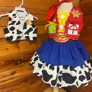 May include: A child's costume featuring a cow print vest with a gold star, and a dress with a blue skirt and a cow print layer. The costume includes a red vest with a gold star and gift box details, and a red neckerchief.