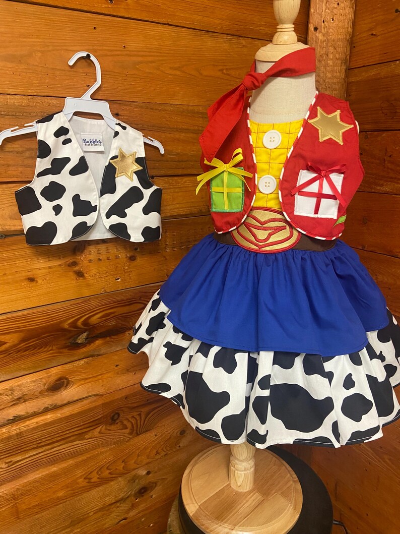 May include: A child's costume featuring a cow print vest with a gold star, and a dress with a blue skirt and a cow print layer. The costume includes a red vest with a gold star and gift box details, and a red neckerchief.