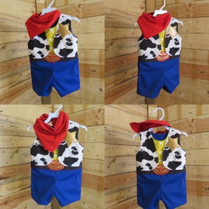 May include: A cowboy-themed costume for a child, featuring a vest with a cowhide pattern, a yellow shirt, blue shorts, and a red bandana. The costume is displayed on a white hanger against a wooden background.