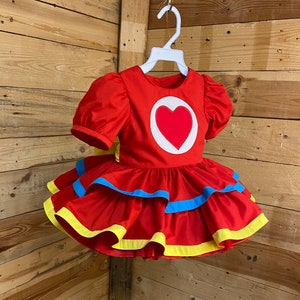 Pode incluir: Vestido vermelho com um grande cora&ccedil;&atilde;o na frente. O vestido tem uma saia rodada com acabamento amarelo e azul.