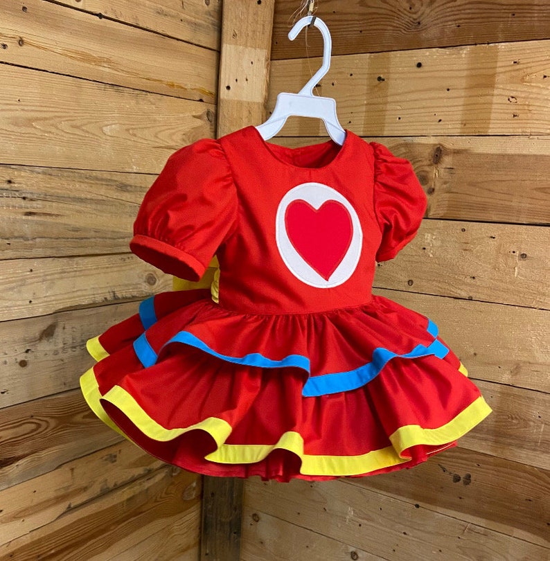 Pode incluir: Vestido vermelho com um grande cora&ccedil;&atilde;o na frente. O vestido tem uma saia rodada com acabamento amarelo e azul.